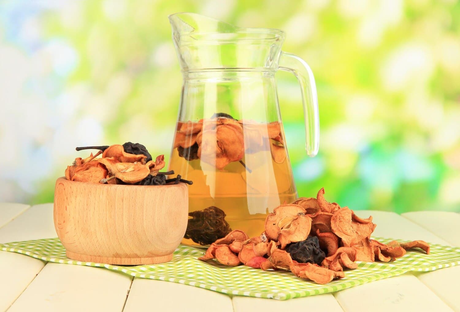 Carafă de sticlă cu compot gălbui în care plutesc felii de mere uscate și prune, lângă un bol de lemn și o grămadă de mere și pere uscate, pe un ștergar în carouri verzi