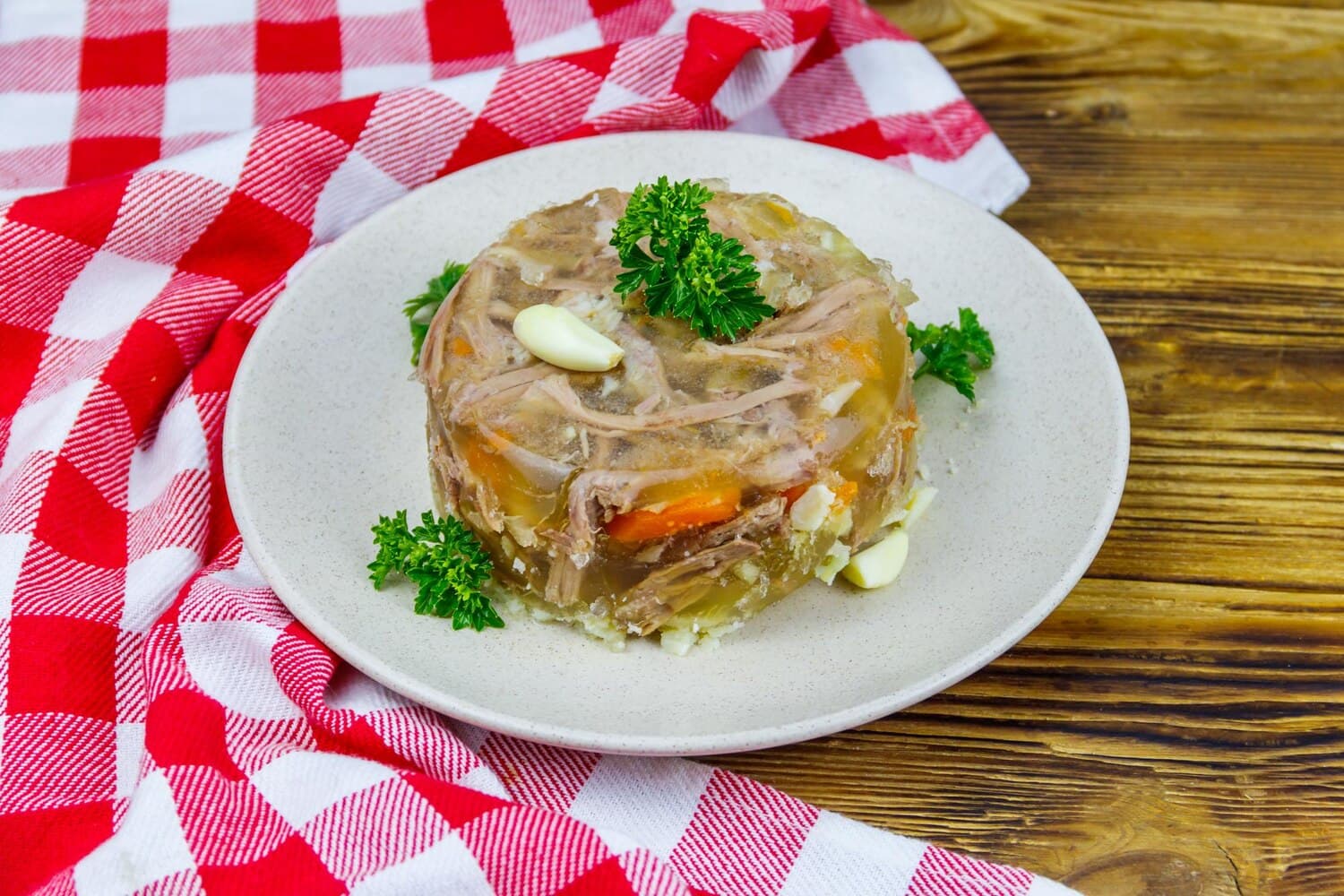 Porție de răcitură răsturnată pe o farfurie, cu bucăți de carne și felii de morcov prinse în zeama încheiată, căței de usturoi și frunze de pătrunjel, pe un ștergar în carouri roșii