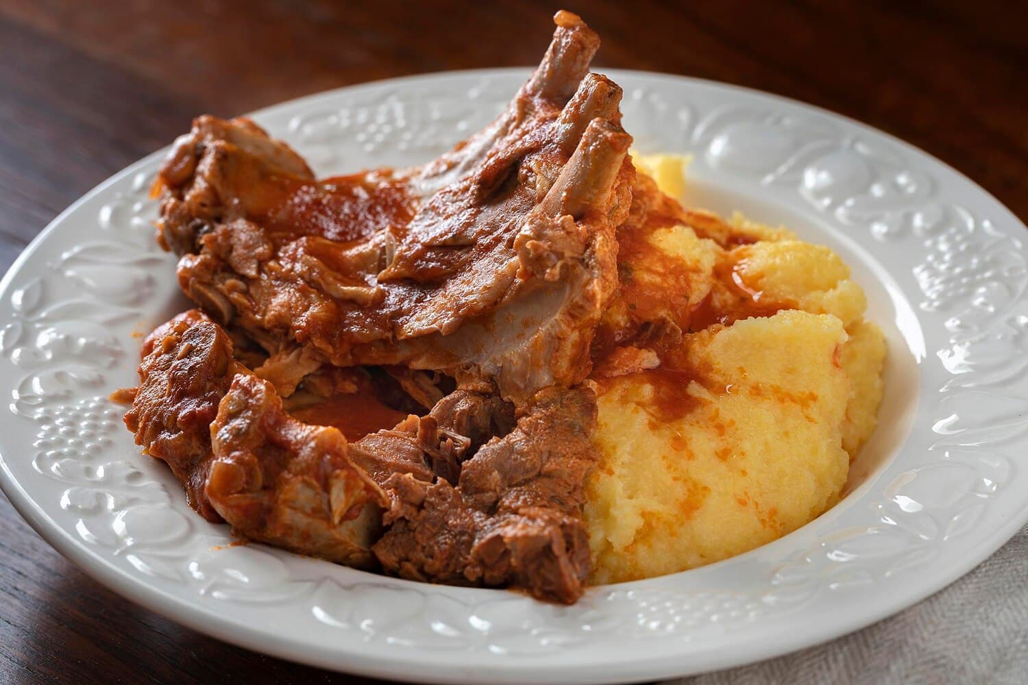 Coaste și bucăți de carne de porc înăbușite în sos de roșii, lângă mămăligă, într-o farfurie albă cu margine în relief