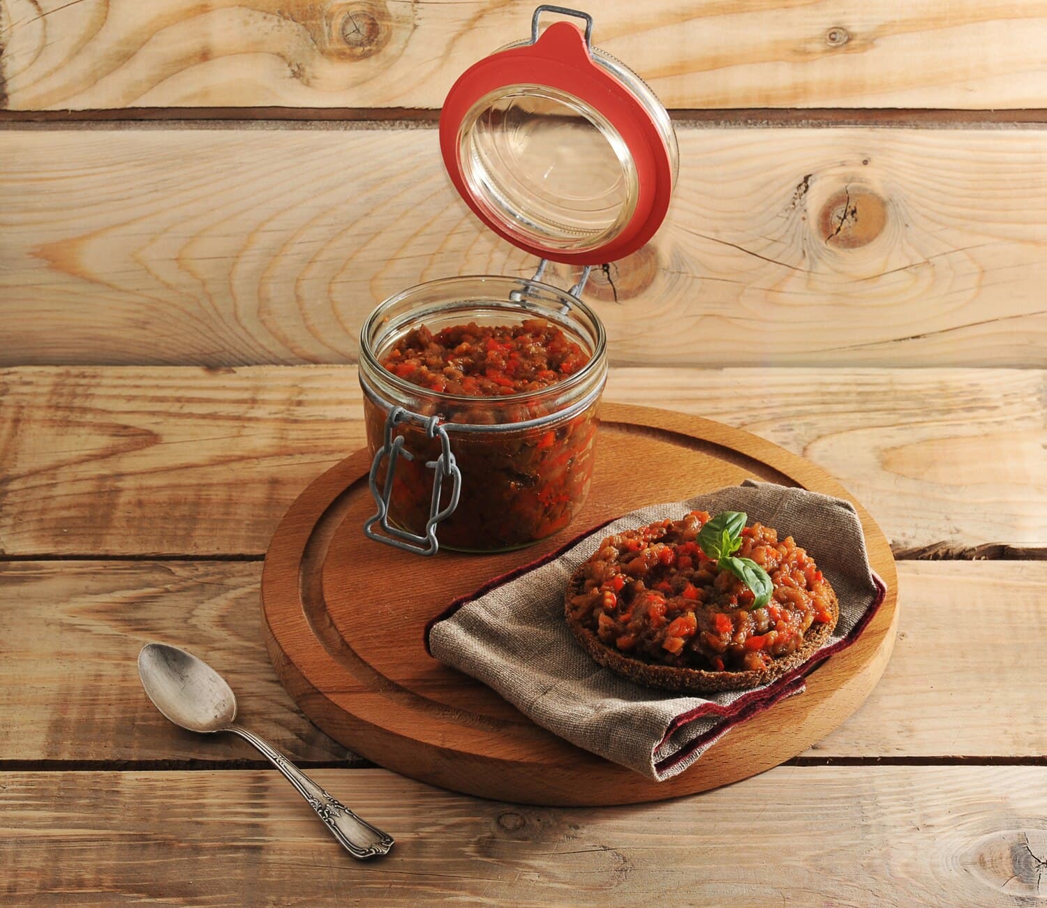 Стеклянная банка с откидной крышкой, полная закуски из рубленых баклажанов и красного перца, и ломтик чёрного хлеба с закуской и листиком базилика на деревянной доске